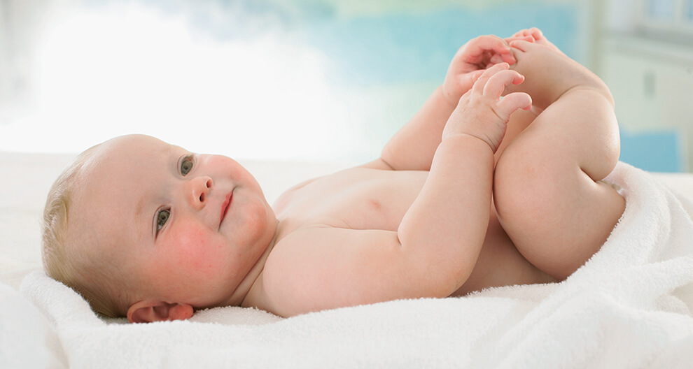 A baby lies on a white blanket, holding its foot.