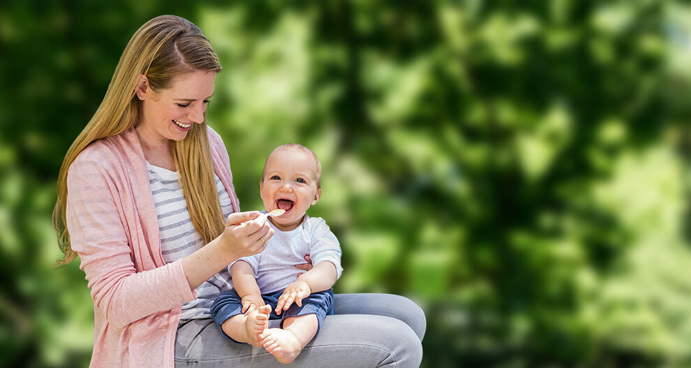 A woman in a pink cardigan and gray pants is feeding a baby with a spoon.