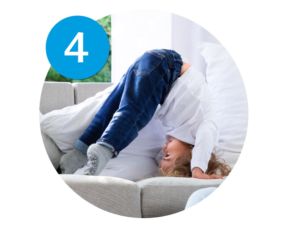 A child is bending forward on a couch, with their head down, wearing a white shirt and blue jeans.