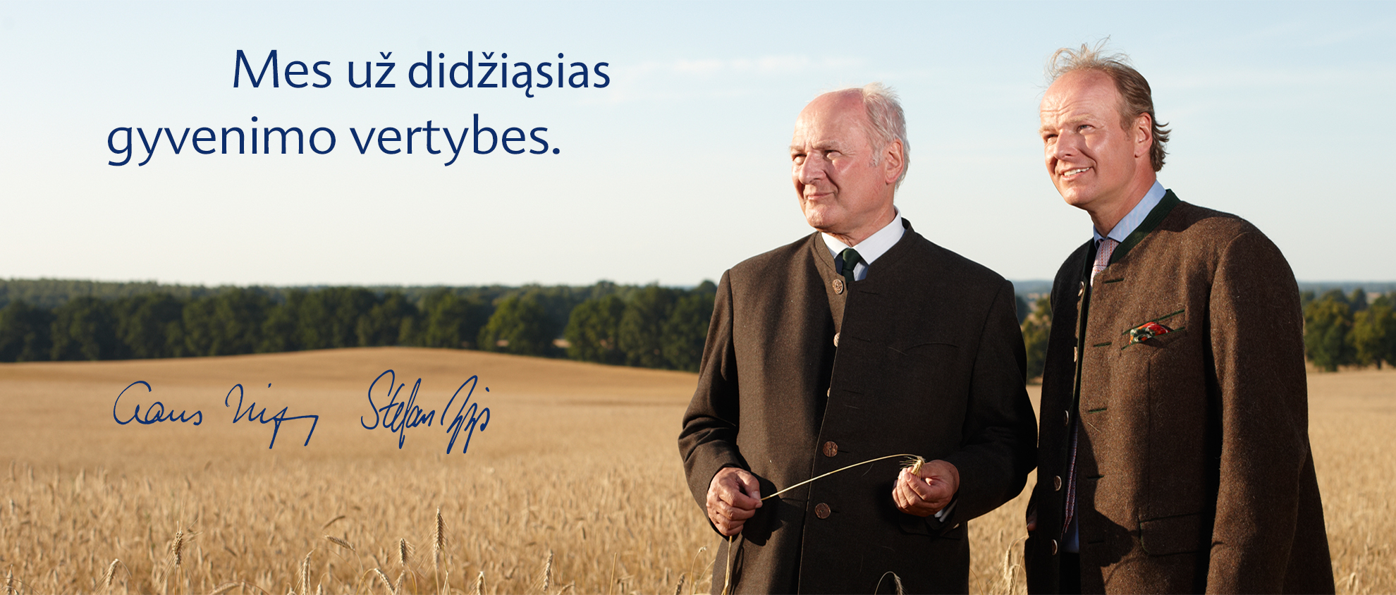 Du kostiumais vilkintys vyrai stovi kviečių lauke, kurio fone matosi medžiai ir giedras dangus.