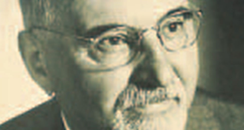 Stambiu planu matomas vyro veidas, kuriame matyti akys, nosis ir burna, vyras su barzda ir ūsais, dėvi akinius.