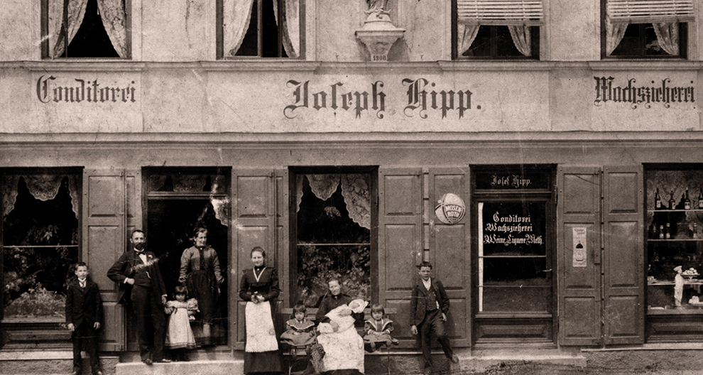 Prie parduotuvės su užrašu "Johann Hipp" stovi ir sėdi grupė žmonių, įskaitant suaugusiuosius ir vaikus.