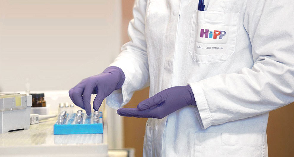Asmuo baltu laboratoriniu chalatu su logotipu "HiPP" ir pavadinimu "CH. OBERMAIER" tvarko mėlyną padėklą su smulkiais daiktais, galbūt laboratorijoje.
