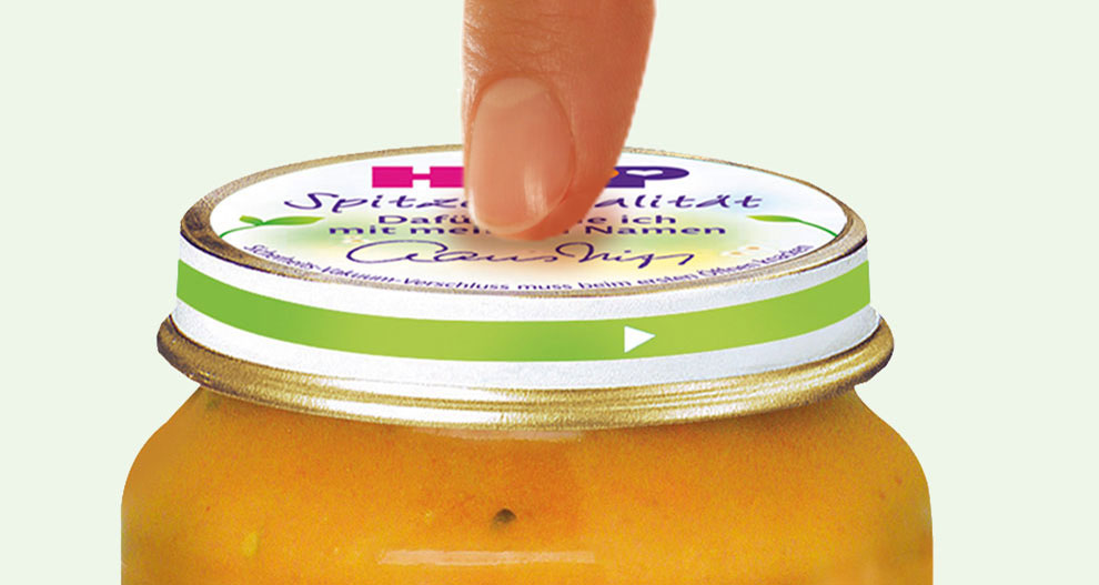 Ranka ketina atidaryti oranžinės spalvos maisto produkto stiklainį su žaliu dangteliu.