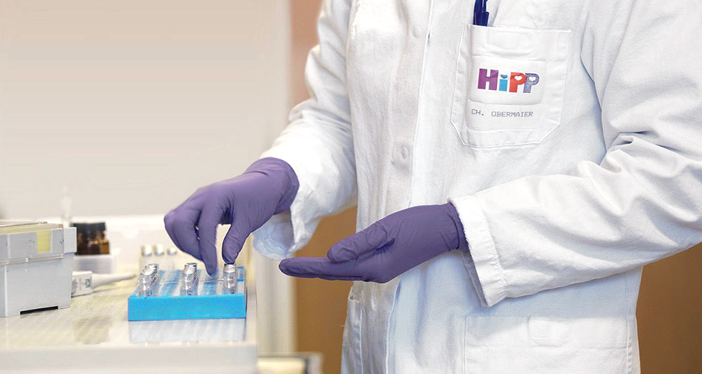 Asmuo baltu laboratoriniu chalatu su logotipu "HiPP" ir pavadinimu "CH. OBERMAIER" tvarko mėlyną padėklą su mažais buteliukais.